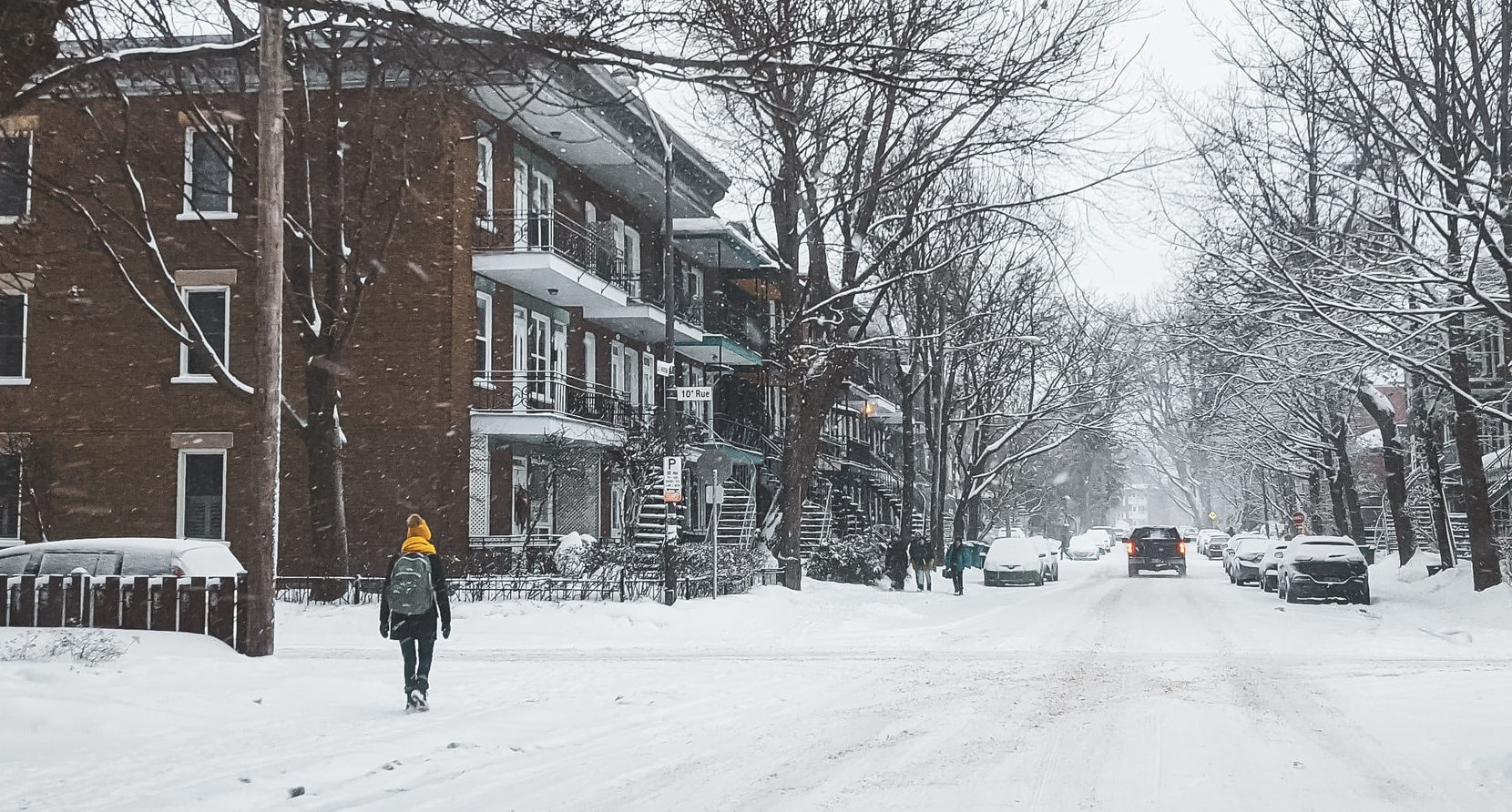A child wearing a backpack walks down a snowy city neighborhood street