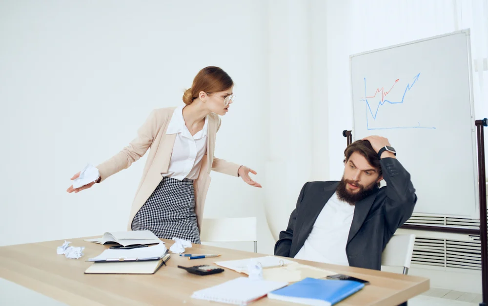 Standing woman with arms outstretched in a questioning stance apparently upset at her seated male coworker who is looking away and is resting a hand on top of his head.