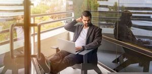 man sitting on gray arm chair using silver laptop computer on building balcony at daytime